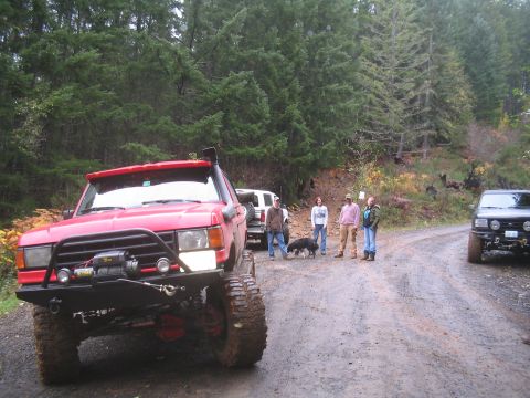 1989 Ford Bronco - A group of people and dogs stand on a dirt road next to a lifted red SUV with off-road modifications.