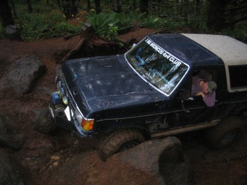 1989 Ford Bronco - A black Ford Bronco with a tan top is shown off-roading through mud and rocks, with a driver visible.