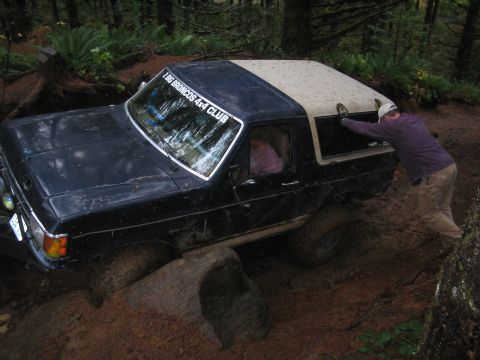 1989 Ford Bronco - A dark blue Ford Bronco with a tan top is stuck in mud on a wooded trail, with a person pushing from behind.