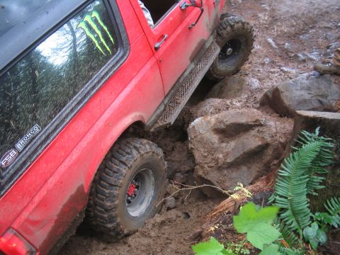 1989 Ford Bronco - A red 1989 Ford Bronco tackles a challenging off-road course, navigating through mud and large rocks.