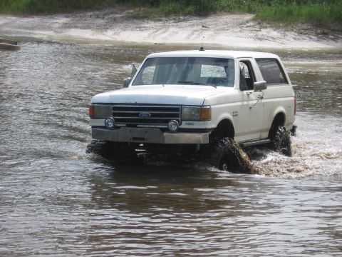 1989 Ford Bronco - A white Ford Bronco navigates through a shallow body of water, kicking up mud.