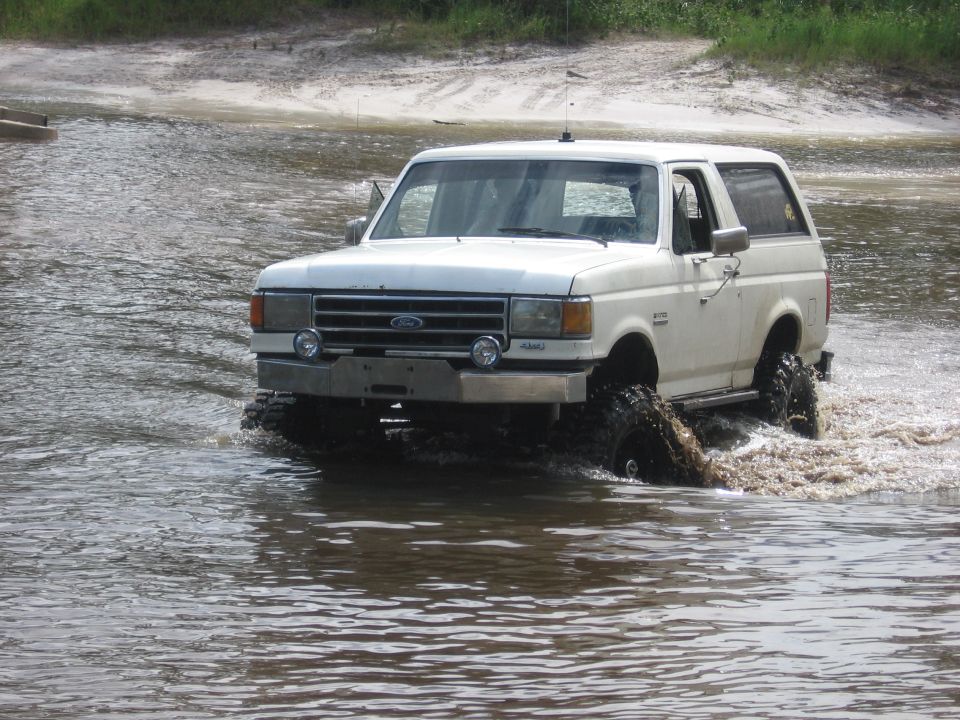 White Ford Bronco with large tires driving through muddy water, front bumper visible