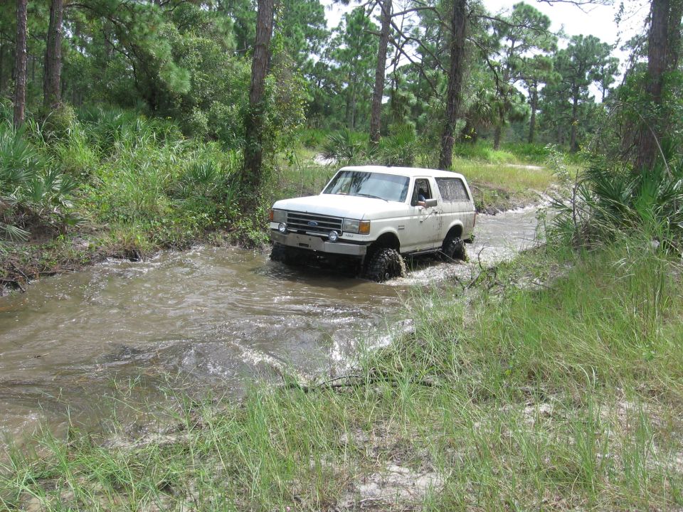 1989 Ford Bronco driving through a muddy stream in a wooded area