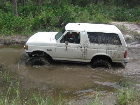 1989 Ford Bronco - A white Ford Bronco navigates through a water-filled trail, kicking up mud.