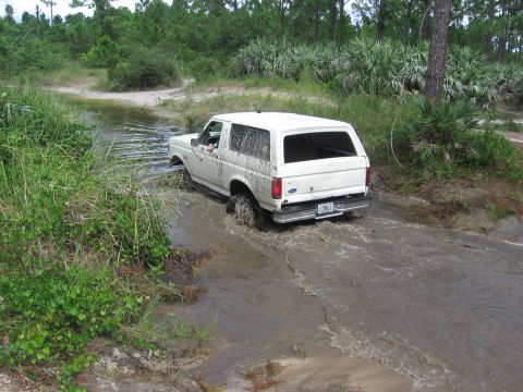 1989 Ford Bronco - A white Ford Bronco navigates a muddy water crossing in a wooded area.