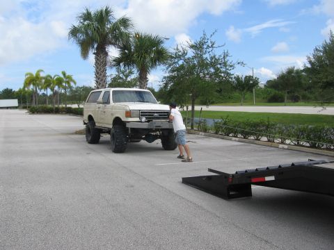1989 Ford Bronco - A person pushes a lifted white Ford Bronco towards a trailer in a parking lot with palm trees.