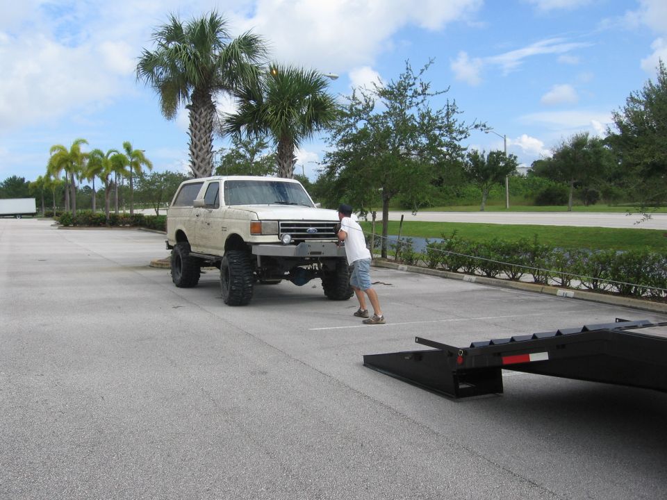 White lifted Ford Bronco being pushed toward a trailer. Palm trees in background