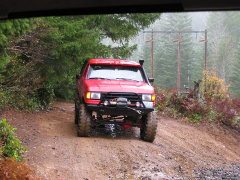 1979 Ford Bronco - A red SUV with off-road modifications drives on a wet, muddy dirt road through a forest.