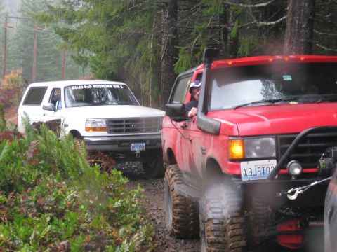 1979 Ford Bronco - Two off-road SUVs navigate a muddy, wooded track during an event.