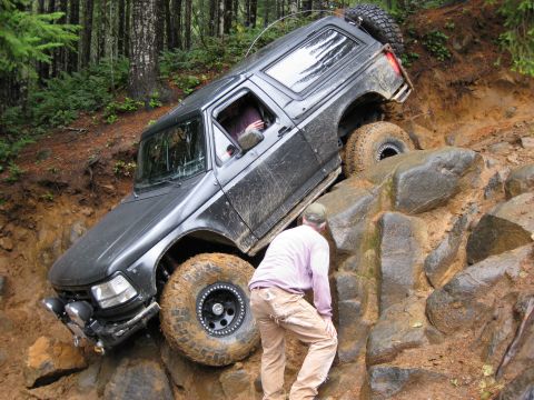 1979 Ford Bronco - A dark SUV navigates a challenging rocky off-road course with a person assisting on the side.