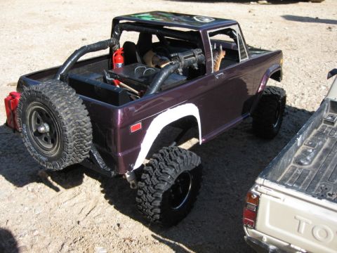 1988 Ford Bronco - A detailed remote-controlled Bronco sits beside a pickup truck in a sandy outdoor area.