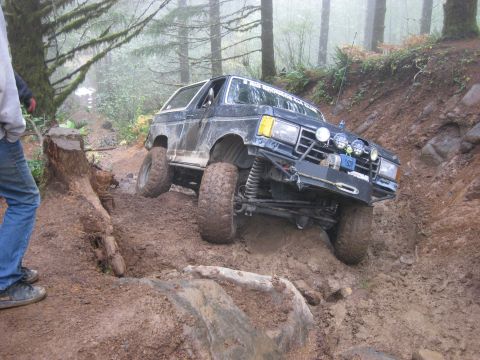 1979 Ford Bronco - A modified black SUV navigates a challenging muddy off-road course in a forest.
