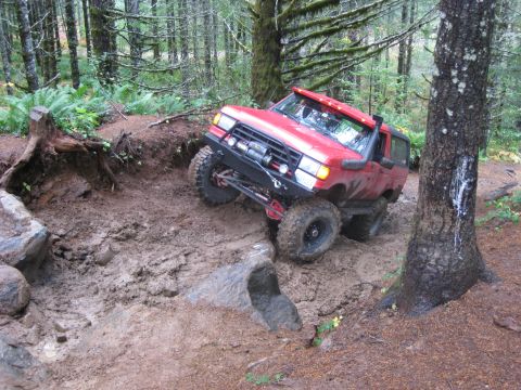 1979 Ford Bronco - A red SUV navigates a challenging muddy trail in a forest setting, demonstrating its off-road capabilities.