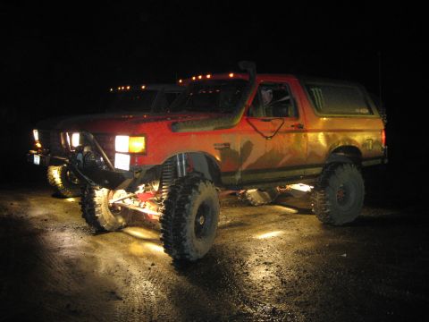 1979 Ford Bronco - A modified red SUV with off-road tires is parked on a dirt road at night, illuminated by its own lights.