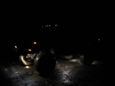 1979 Ford Bronco - A dark, off-road vehicle with large tires is parked on a wet surface at night, illuminated by its own lights.