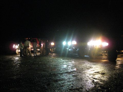 1979 Ford Bronco - Several trucks are parked on a wet road at night, illuminated by their headlights, with a group of people gathered nearby.