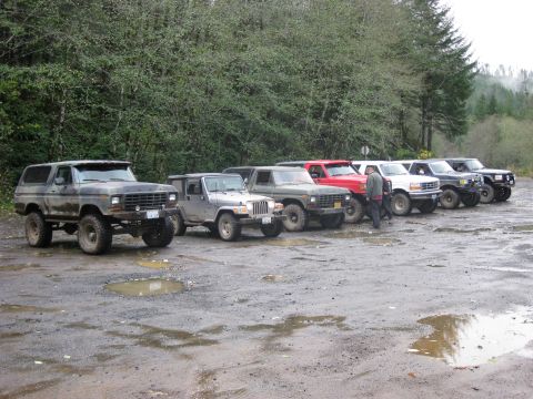 1979 Ford Bronco - A line of off-road vehicles, including a 1979 Ford Bronco, parked on a muddy lot.