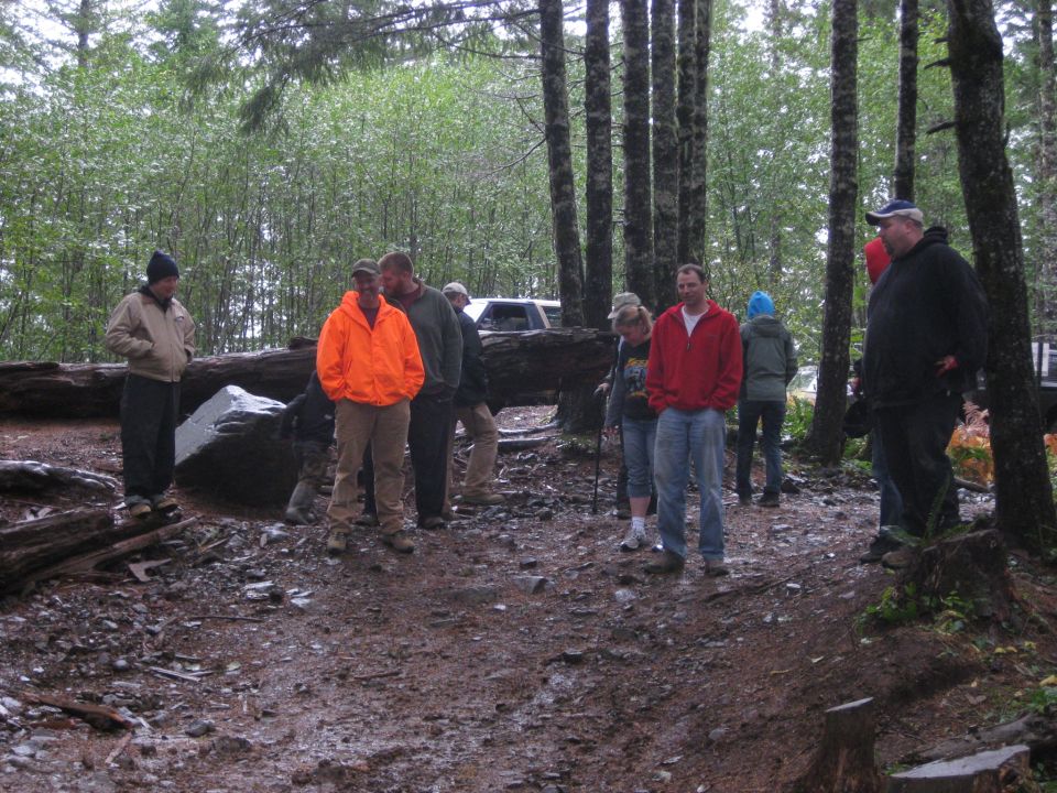 group of people standing around in a forest clearing with a fallen log and some rocks