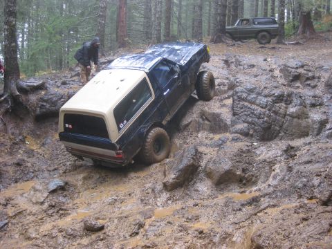 1979 Ford Bronco - Two Ford Broncos tackle a challenging muddy and rocky off-road course in a wooded area.