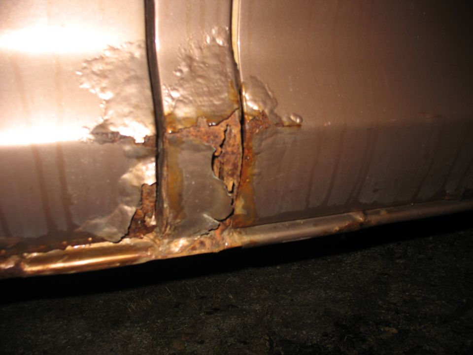 Close-up of rust holes and bubbled paint on lower body panel of tan vehicle at night