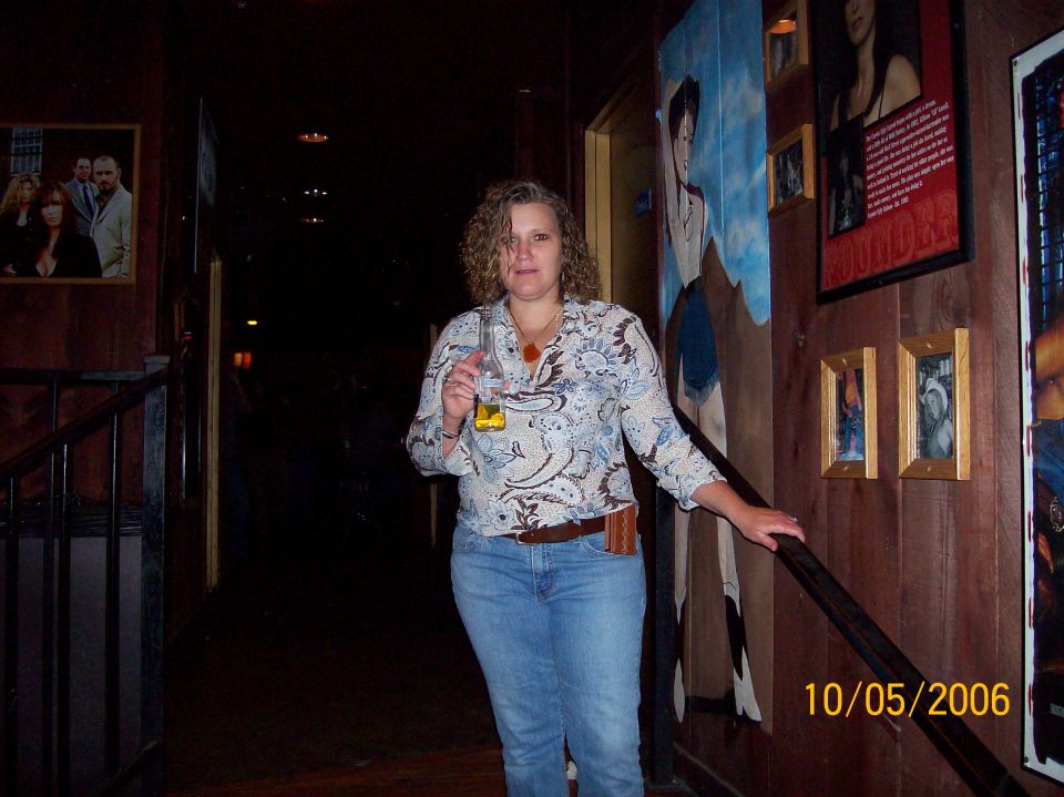 Woman holding a beer bottle in a dark bar hallway with framed pictures on wood walls, dated 10/05/2006