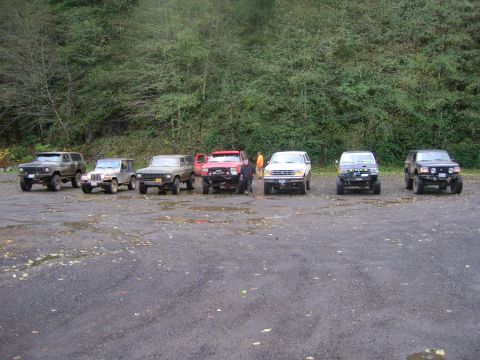 1996 Ford Bronco - A group of off-road vehicles are parked in a lot with a wooded area in the background.