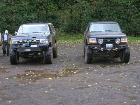 1996 Ford Bronco - Two modified Ford Broncos are parked side-by-side on a wet, leaf-strewn surface with trees in the background.