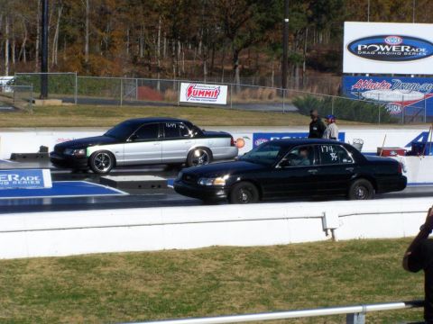 2001 Ford Crown Victoria - Two Ford Crown Victorias compete on a drag strip, with racing banners in the background.