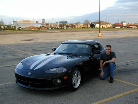 1996 Dodge Viper - A man poses next to a black Dodge Viper with racing stripes in an empty parking lot.