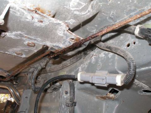 1997 Ford Taurus - Underbody view of a heavily rusted steel brake line running along the floor pan, held by a clip at a corroded bracket. A gray two-pin elect…