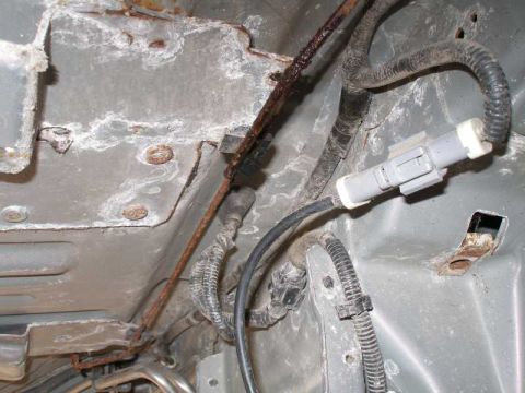 1997 Ford Taurus - Close-up of a vehicle's undercarriage showing a heavily rusted metal brake line running along a dusty gray body panel, with black wiring ha…