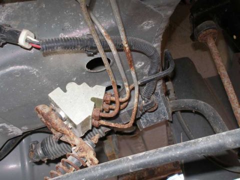 1997 Ford Taurus - Close-up underbody view showing heavily rusted steel brake lines bending near a corroded distribution block or master cylinder area, with b…