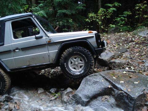 1986 Ford Bronco - A Mercedes-Benz G-Wagen navigates a rocky stream bed in a wooded area.
