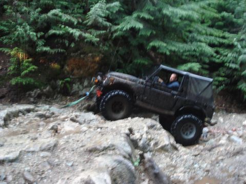1986 Ford Bronco - A dark off-road vehicle with a soft top is being winched through a rocky, wooded terrain.