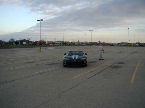 1996 Dodge Viper - A person stands near a black Dodge Viper with white racing stripes in a large, empty parking lot under a cloudy sky.