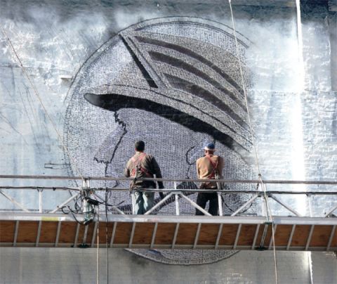 2003 Mercury Marauder - Workers on scaffolding paint a large mural depicting a helmeted head on a textured wall.