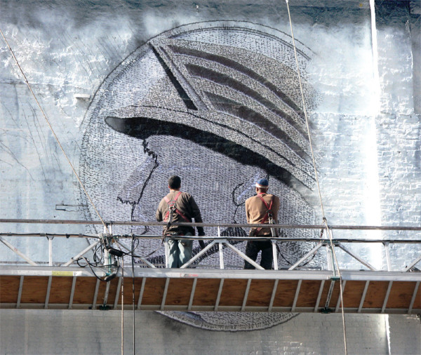 Two workers on scaffolding painting a large mural of a helmeted head