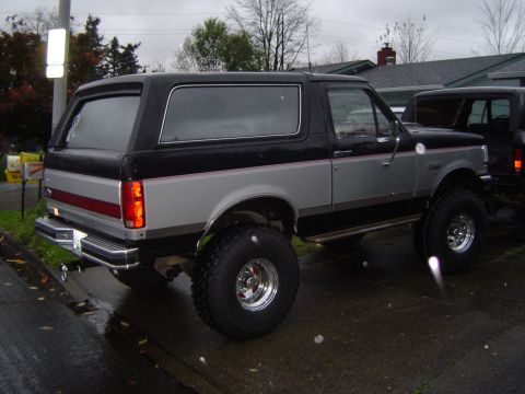 1989 Ford Bronco - This lifted SUV, likely a 1987 model based on its body style, is parked next to another vehicle on a wet day.