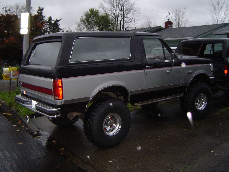 Lifted black and silver Ford Bronco on chrome wheels and tall tires, rear three-quarter view on a wet street