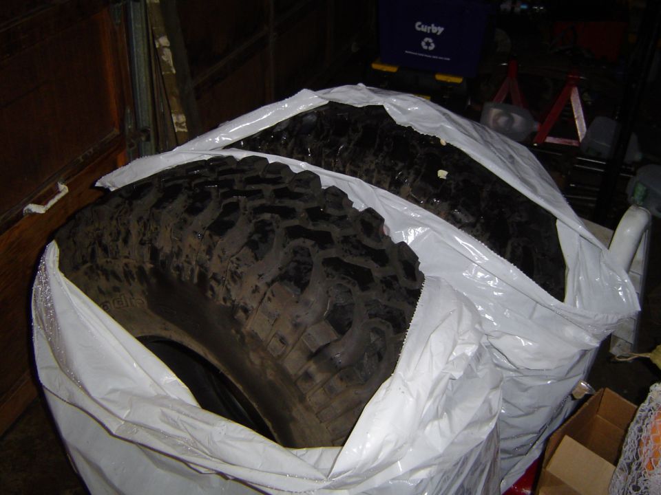 Two dirty mud-terrain tires bagged in white plastic and stored in a dark garage
