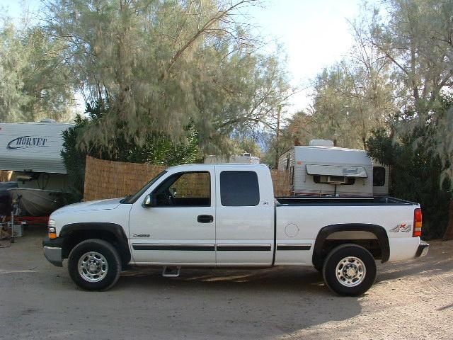 White Chevrolet Silverado extended-cab pickup viewed from the side, parked outdoors near trailers.