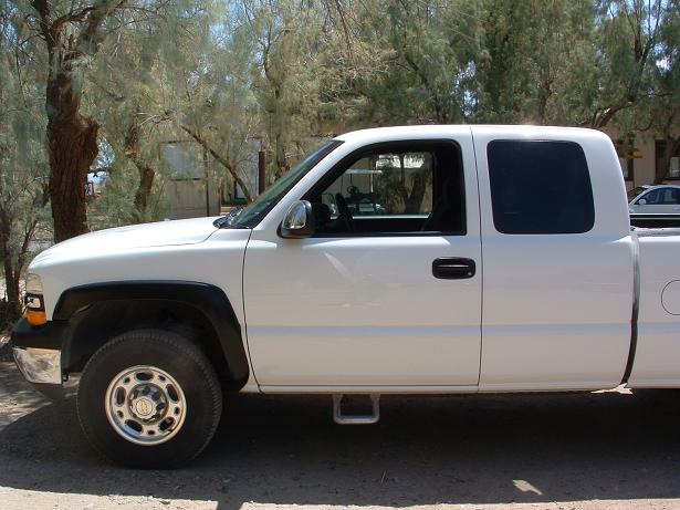 White Chevrolet Silverado extended-cab pickup, side view, parked under trees.