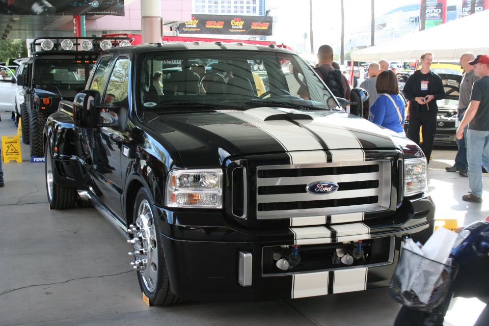 Black Ford F-Series Super Duty truck with white racing stripes, dual rear wheels, and a scoop on the hood. Another vehicle is visible behind it