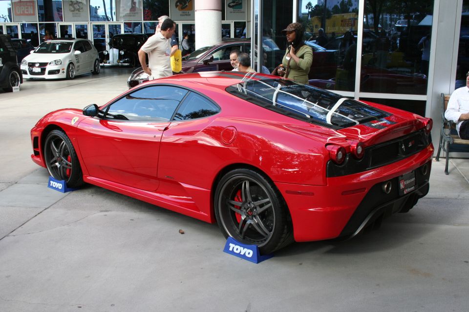 Red Ferrari coupe parked on blue TOYO wheel chocks, with a white Volkswagen Golf and other vehicles in the background