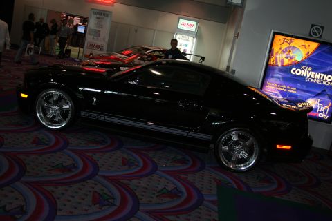 1998 Ford Crown Victoria - A black Ford Mustang Shelby GT500 is displayed indoors with other vehicles and convention signage.