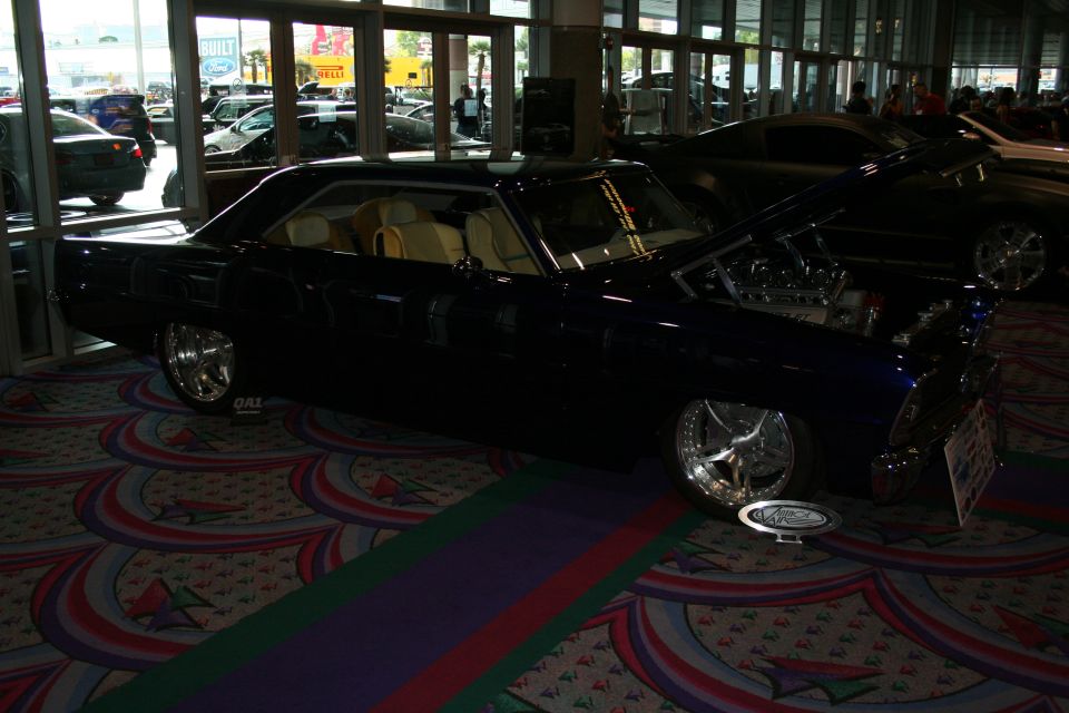 Dark blue coupe with custom wheels and interior, hood open showing engine. Other cars visible in background