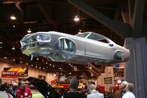 1998 Ford Crown Victoria - A car body is displayed hanging from the ceiling at an automotive show, with spectators gathered below.