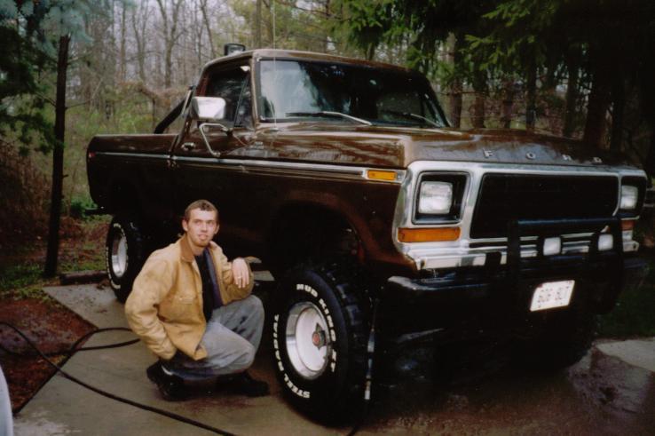 Man kneeling beside lifted brown Ford pickup with white spoke wheels and mud tires on wet driveway