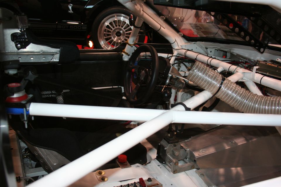 Interior of a racing car with roll cage, racing seat, and steering wheel visible. Another car with "GT 500" text is partially visible in the background