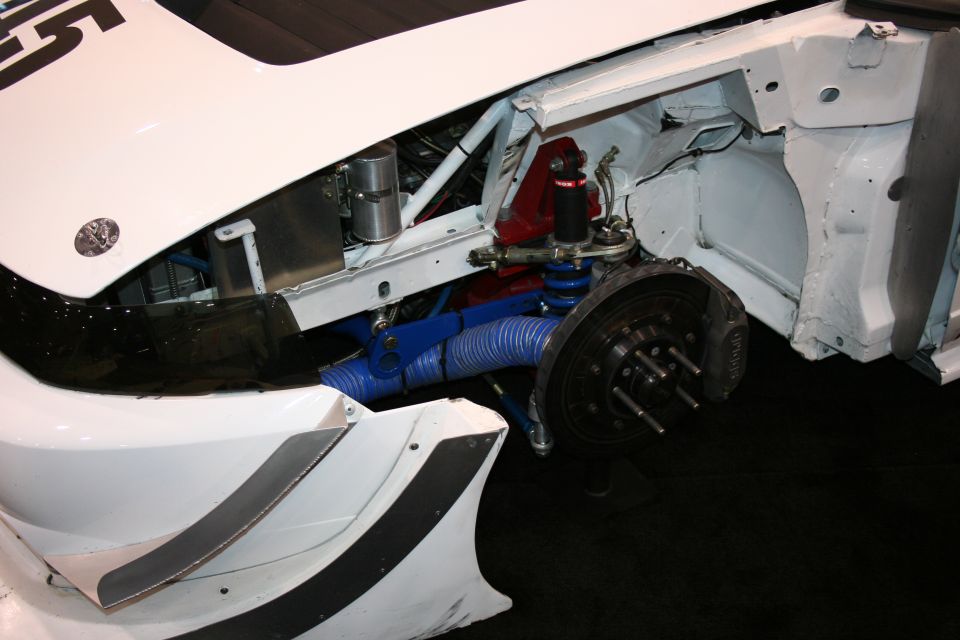 Close-up of a white race car's front suspension, showing blue hoses, red shock, and brake assembly